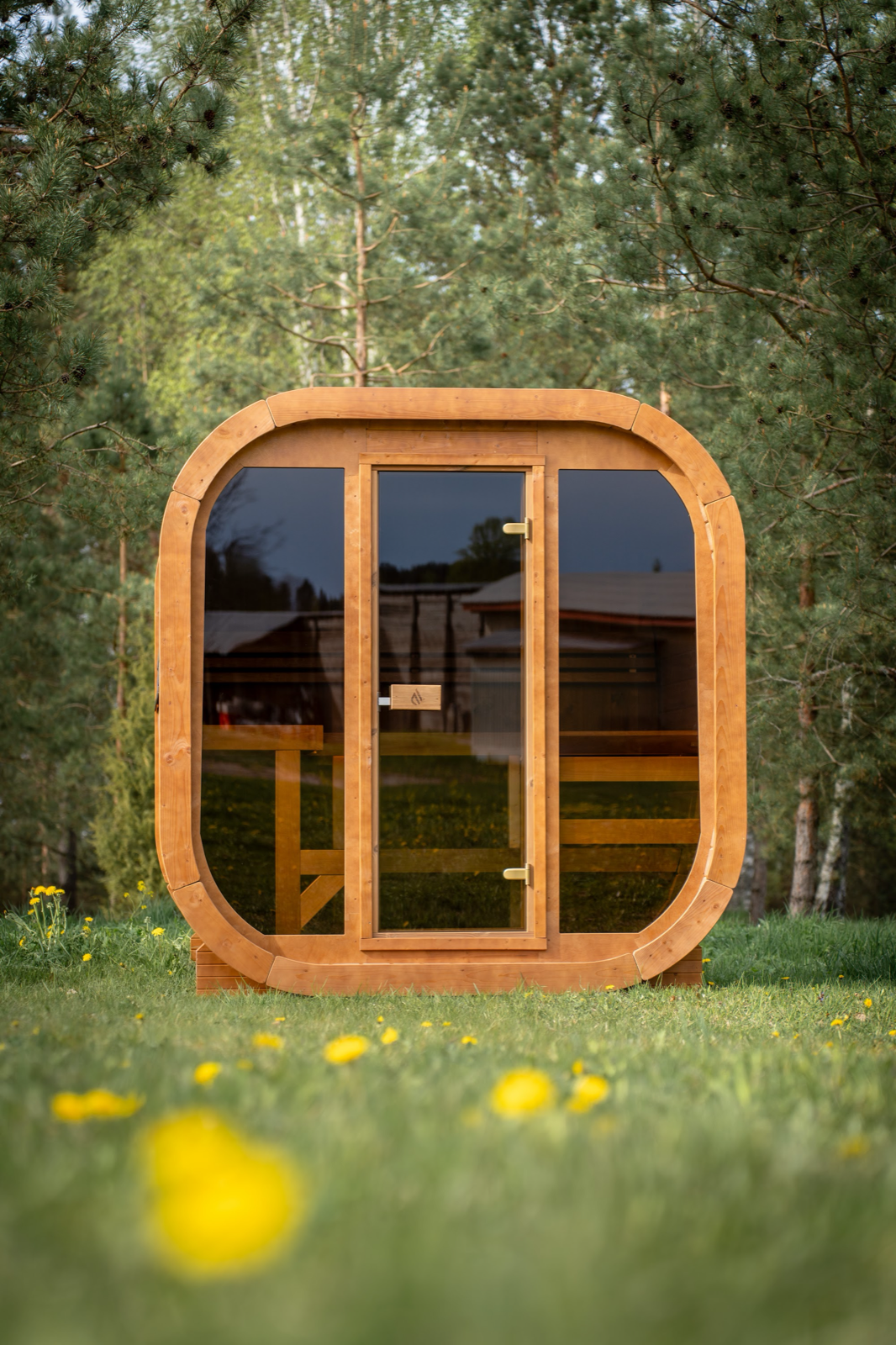 Head-on view of the PlusLife Max Traditional Sauna 2.0 showing timber benches through the full-height glass front, trees reflected in the glass
