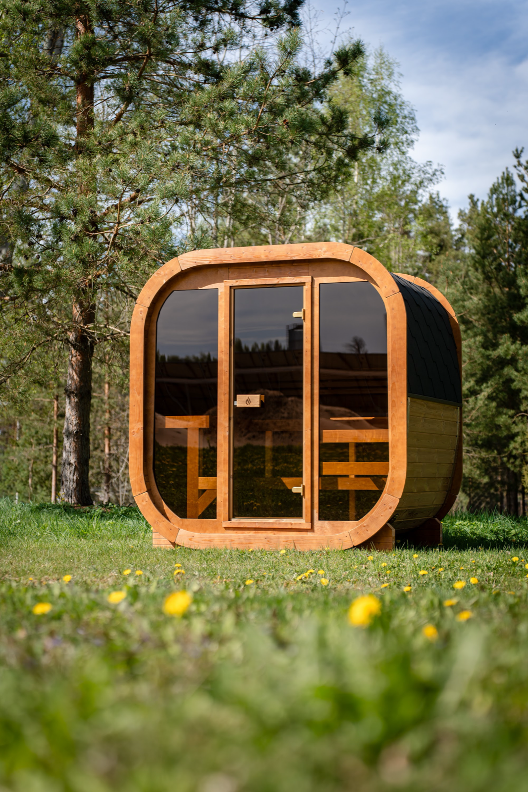PlusLife Max Traditional Sauna 2.0, a rounded-square timber cabin with a full glass front and a dark shingled curved roof, standing on grass in front of pine trees