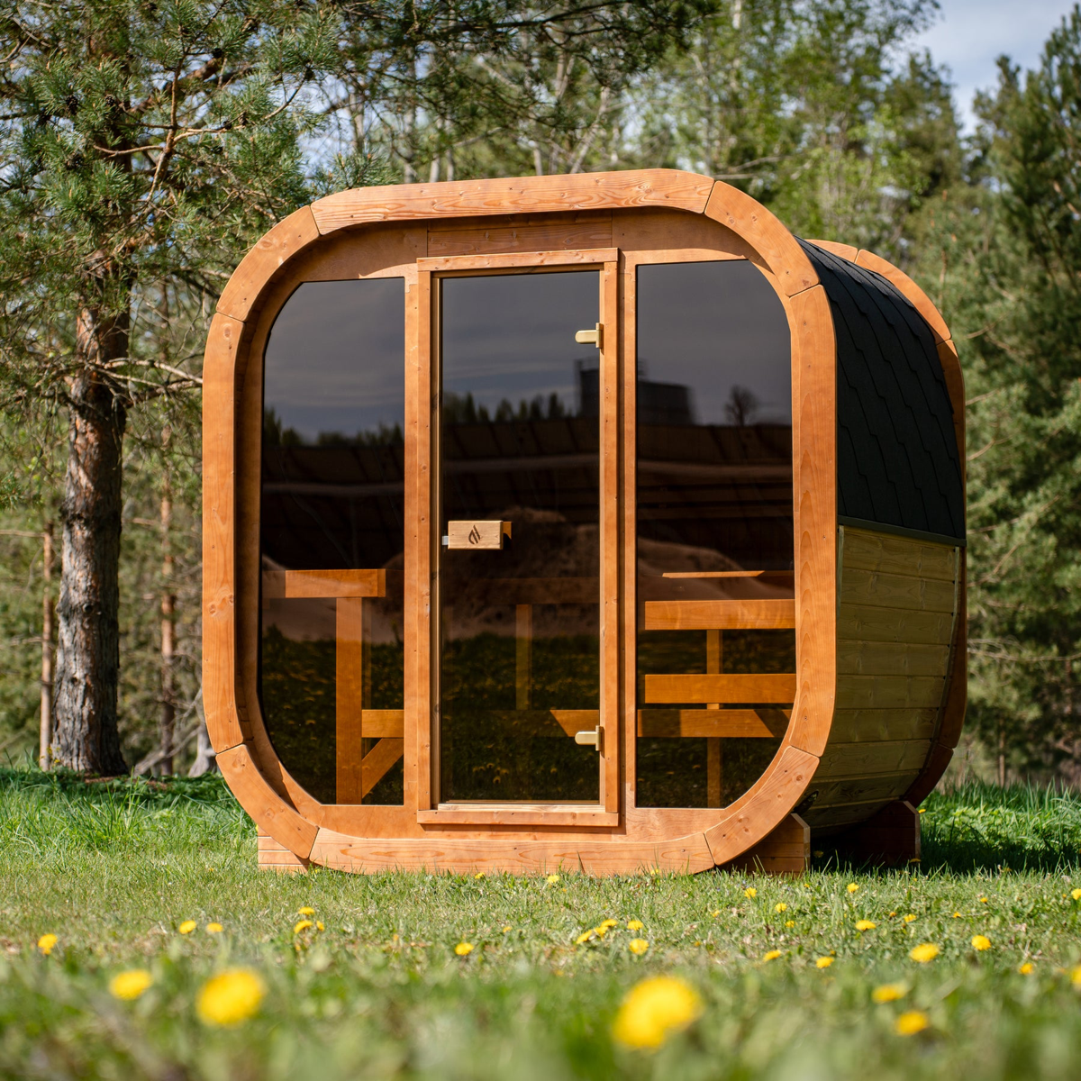 PlusLife Max Traditional Sauna 2.0, an outdoor timber cabin with a rounded timber frame, a full tinted-glass front and a dark shingled roof, photographed outdoors with trees behind it